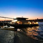 Ferry with lights sailing at dusk near a crowded dock